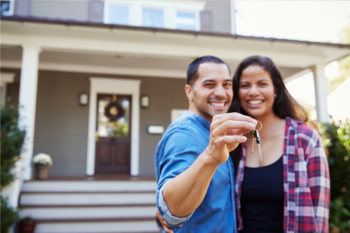 Young couple stands outside first home with keys.
